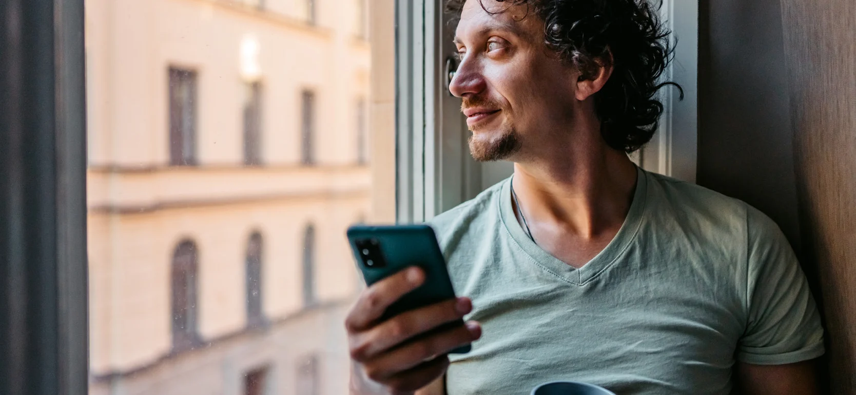 man drinking coffee and using smart phone while leaning on a window sill with Swedish building in the background.