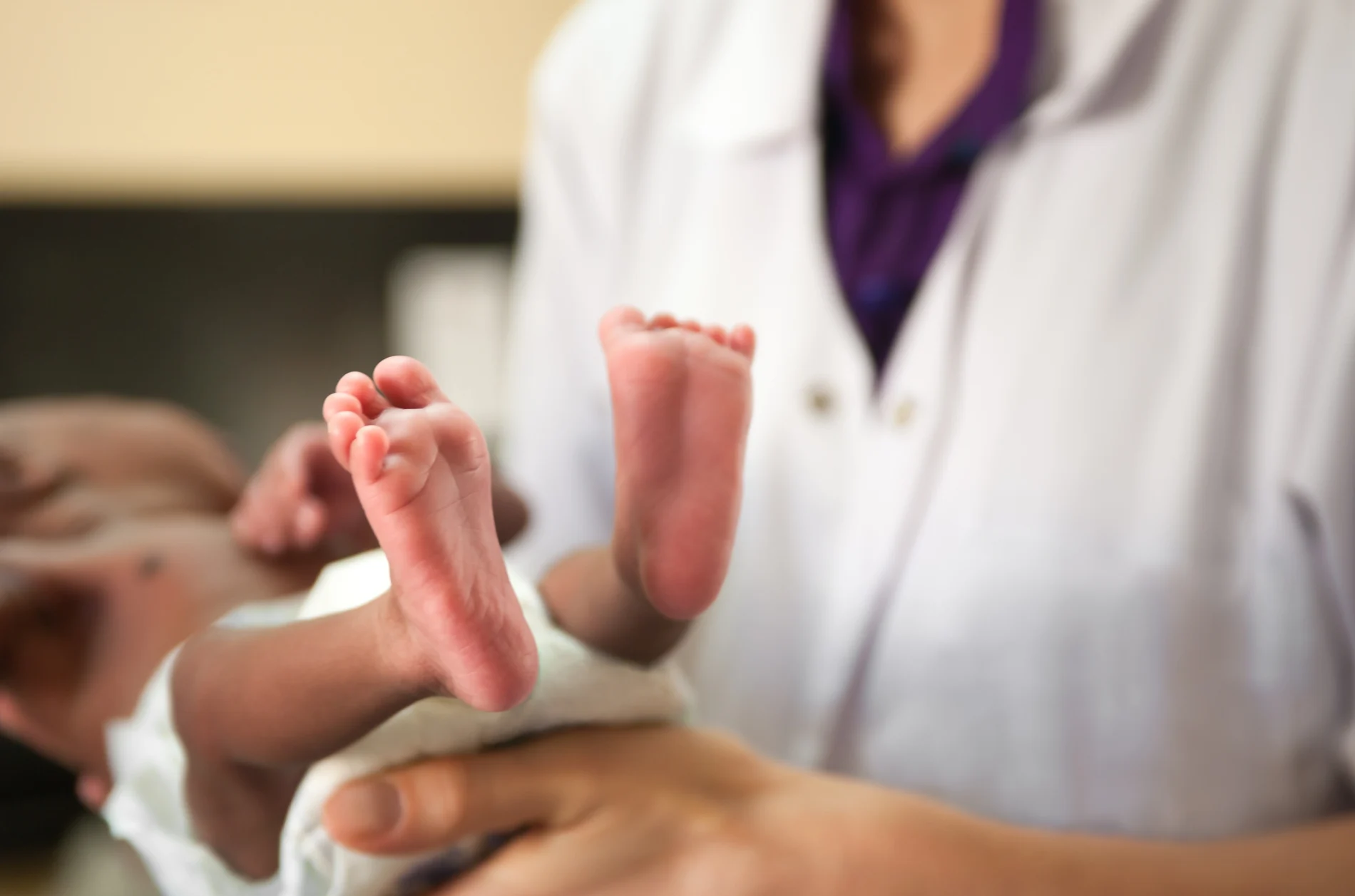 Close-up shot of a premature Black baby being held by a doctor, focused on the baby's feet. The baby is wearing a nappy.