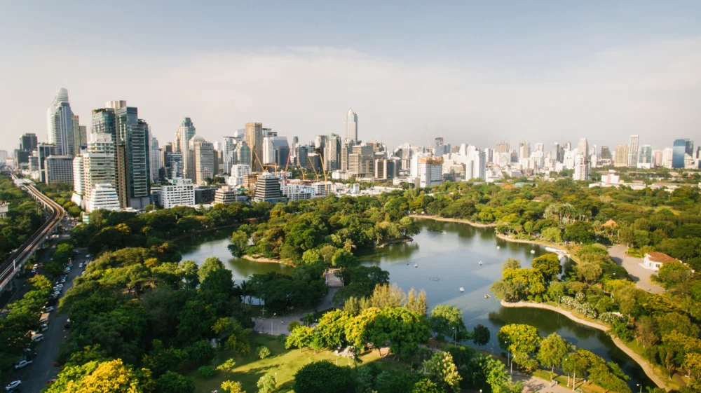 High-angle shot of Lumphini Park, Bangkok. A lake is surrounded by greenery, with a cityscape in the background.