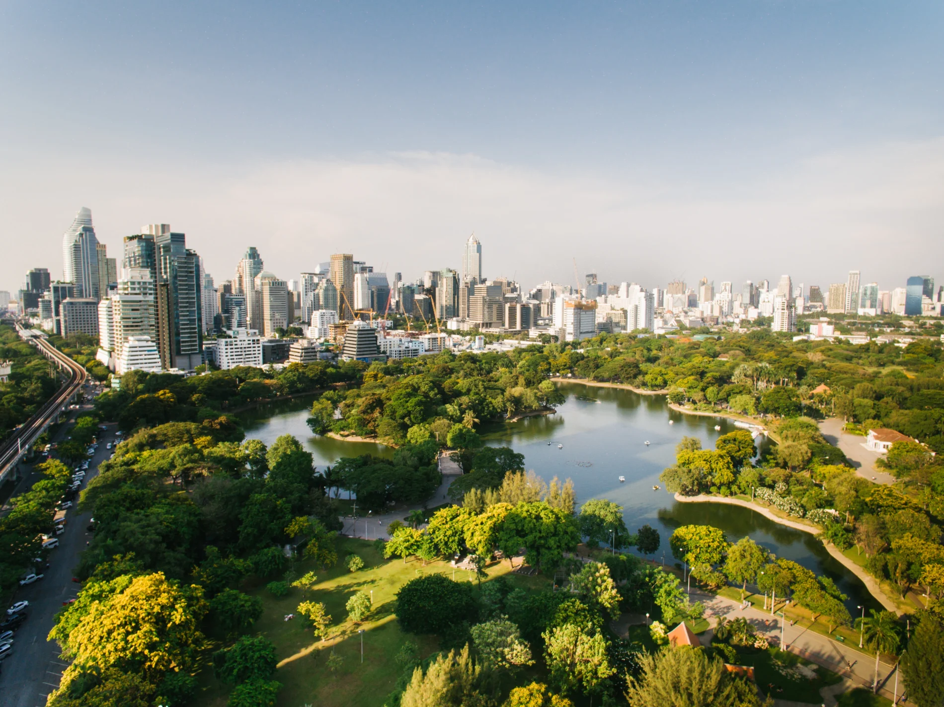 High-angle shot of Lumphini Park, Bangkok. A lake is surrounded by greenery, with a cityscape in the background.