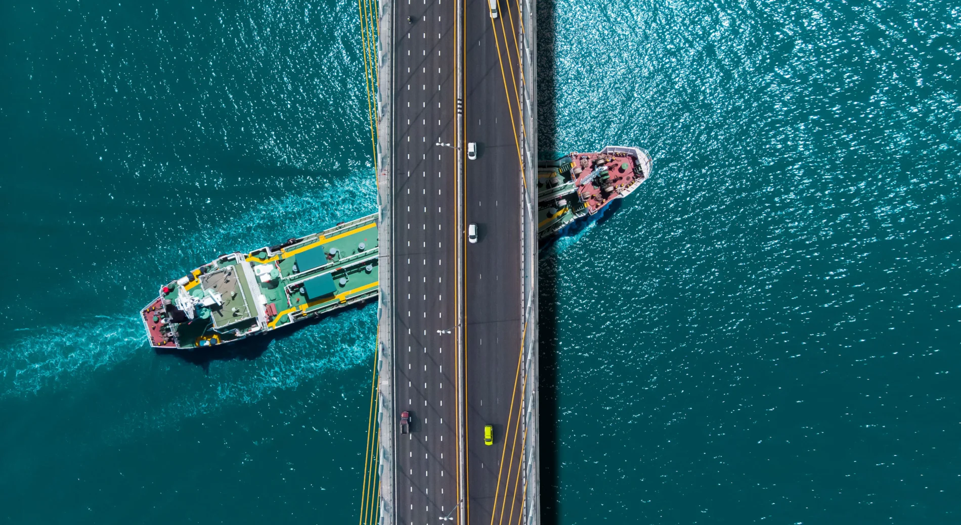 Aerial view over a turquoise sea where an oil tanker passes beneath a sling bridge.