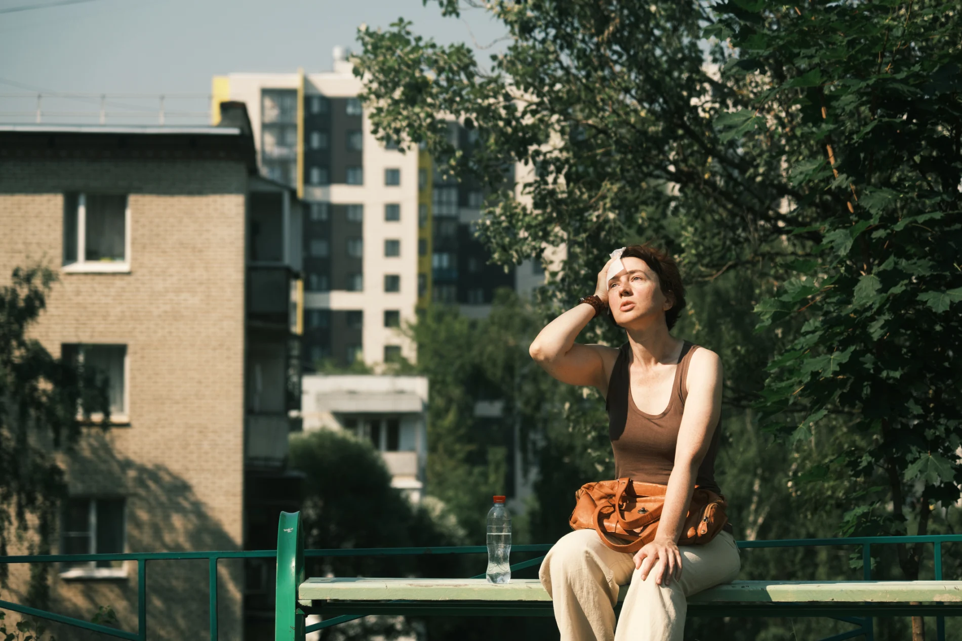 A woman is sitting in the city, buildings and and a tree in the background, visibly feeling bad from heat, holding a hand to her forehead, a watter bottle next to her.