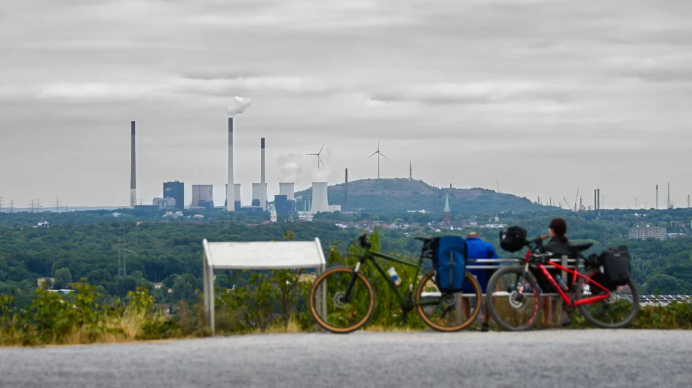 cyclists sit in the foreground on a bench, viewing a green forested landscape with fossil fuel energy, wind turbines, and a town with a church tower in the distance.