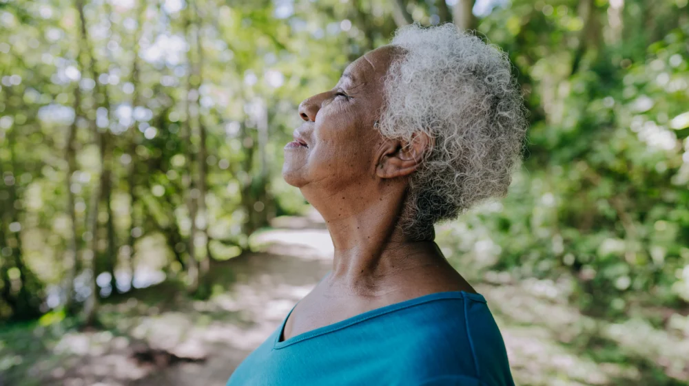 Profile shot of an older Black woman in a forest environment, wearing a blue top, facing left. Her face is turned up towards the sky, eyes closed, as she takes a deep breath.