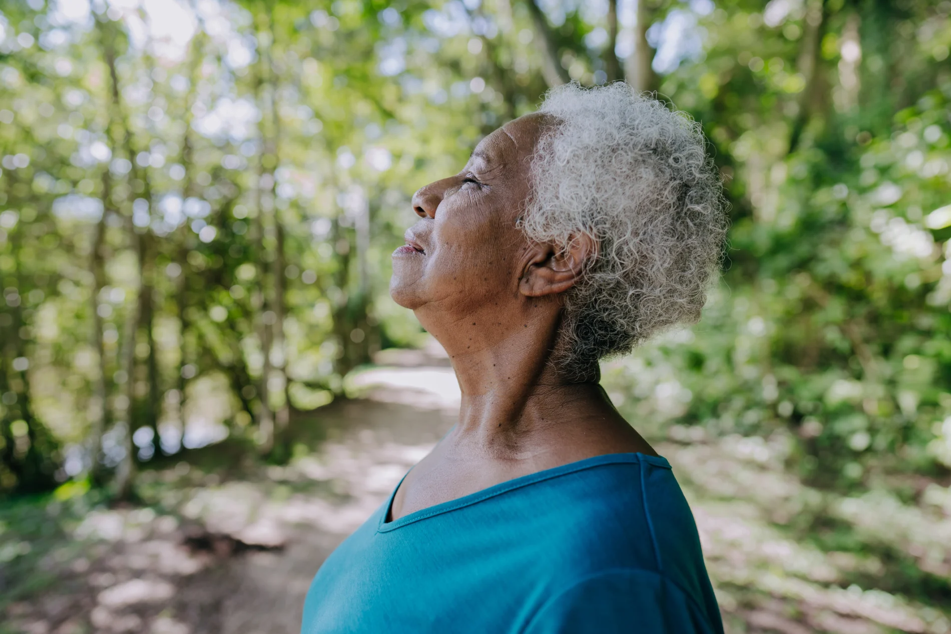 Profile shot of an older Black woman in a forest environment, wearing a blue top, facing left. Her face is turned up towards the sky, eyes closed, as she takes a deep breath.