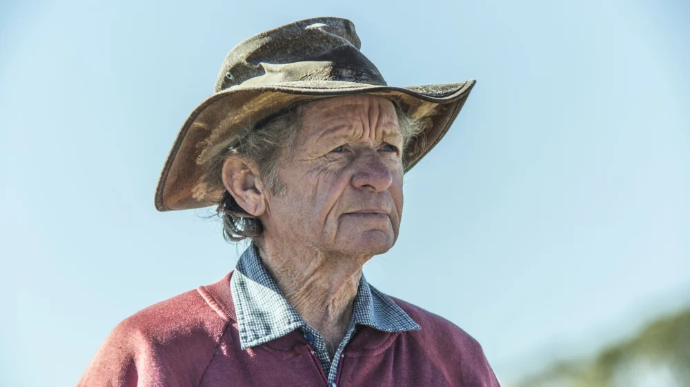 Headshot of older man dressed in outdoor work clothes, wearing a hat, looking into the distance to the right of shot.