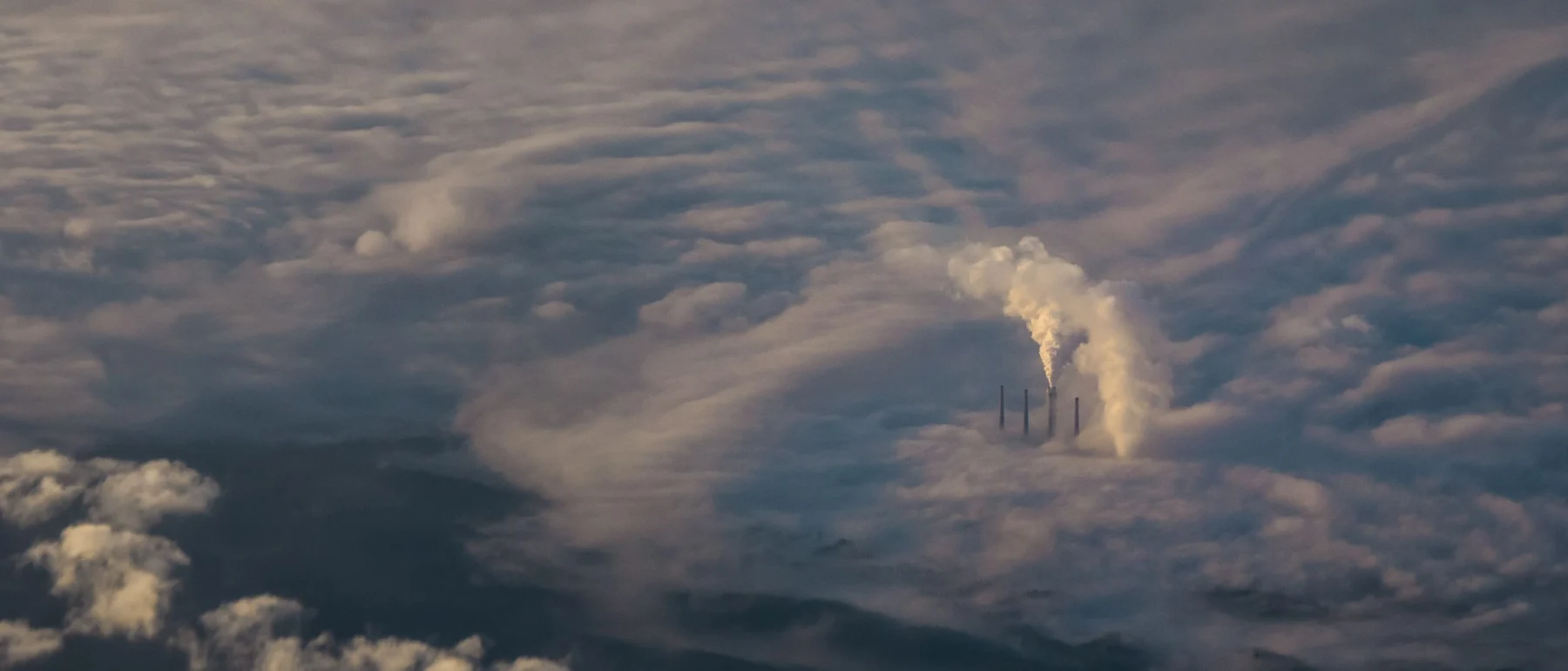 Aerial view of a sea of clouds punctuated with smoke stacks emitting pollution.