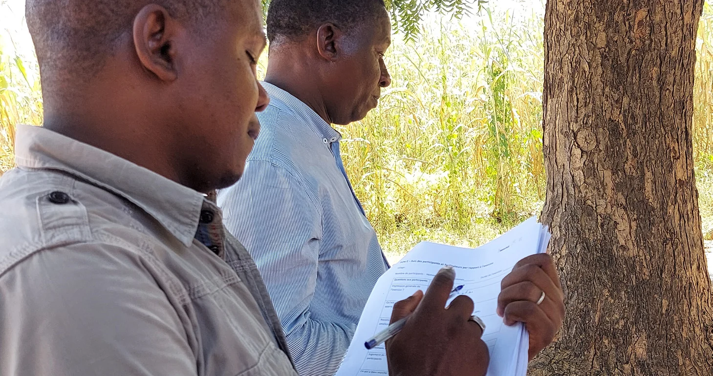 A man in the foreground holds a notebook and is taking notes with a pen.