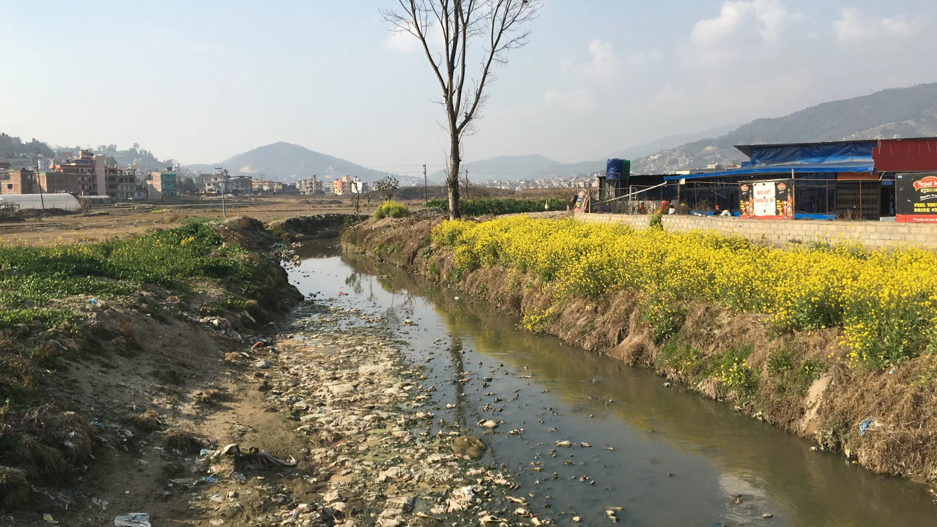 A landscape view of the Manohara informal settlement - Kathmandu, Nepal. There is a waterway in the central foreground, with buildings in the distance either side of it. Himalayas in the background.