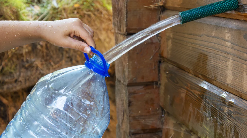 A person's hand holding a blue funnel to direct water from a large plastic bottle into a green hose against a wooden backdrop.