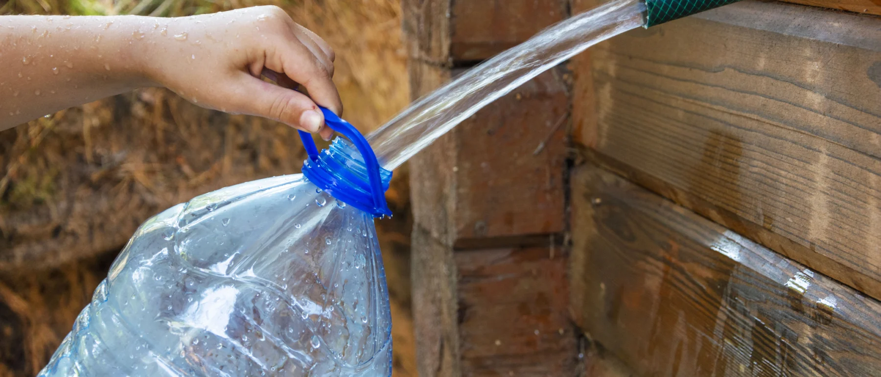 A person's hand holding a blue funnel to direct water from a large plastic bottle into a green hose against a wooden backdrop.