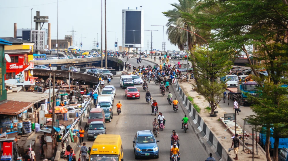 High-angle shot of a road network which extends from the foreground all the way to the horizon. Traffic mostly flows towards the camera: cars, motorbikes, trucks. A bustling street dominates the left of the shot.