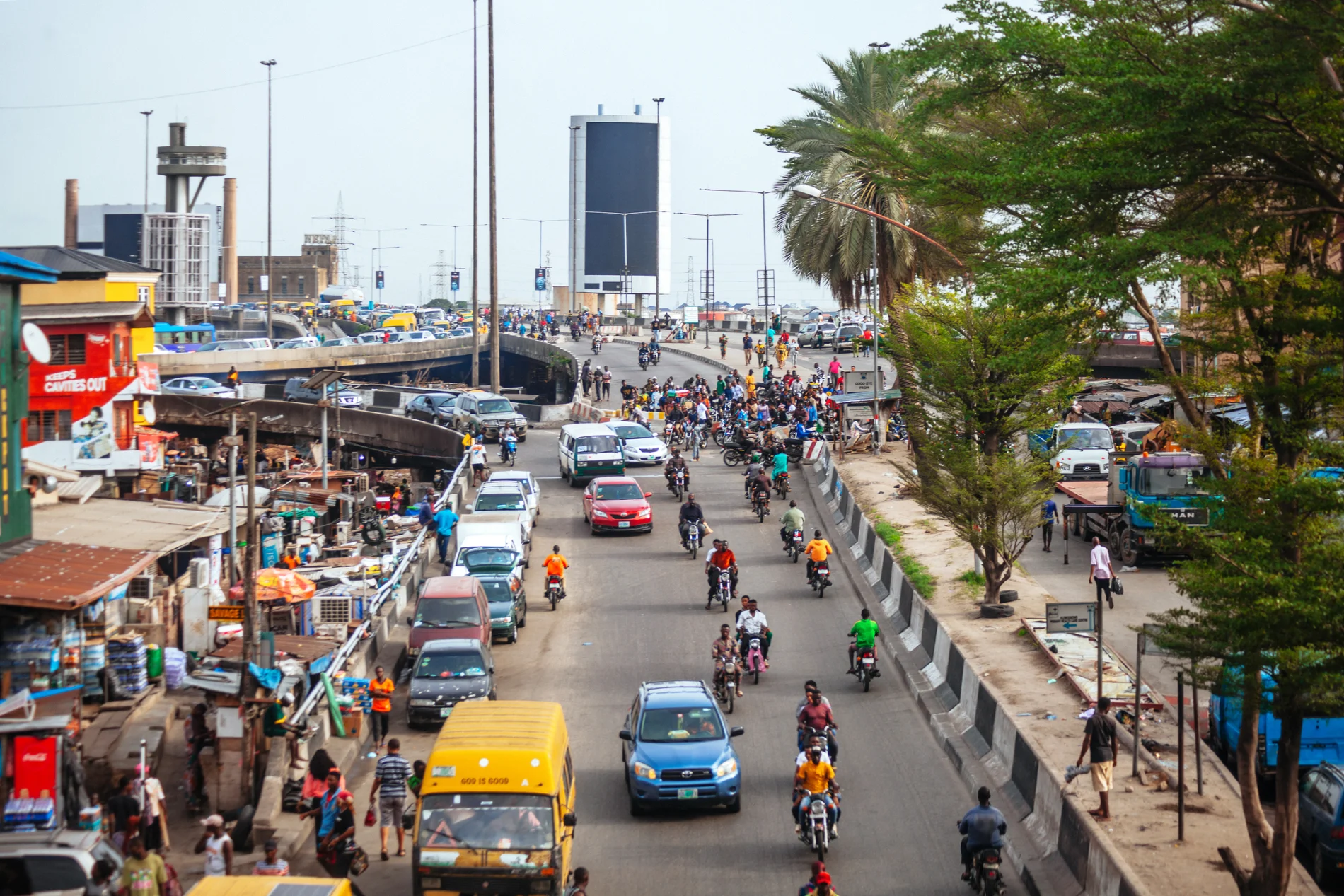 High-angle shot of a road network which extends from the foreground all the way to the horizon. Traffic mostly flows towards the camera: cars, motorbikes, trucks. A bustling street dominates the left of the shot.