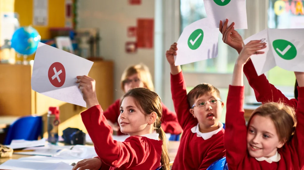 Primary school children in a classroom holding up pieces of paper with green ticks or red crosses.
