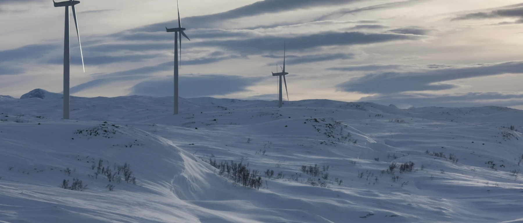 A small windfarm near Bjornafell, Sweden: three turbines atop a snow-covered hill and plain below under cloudy skies.