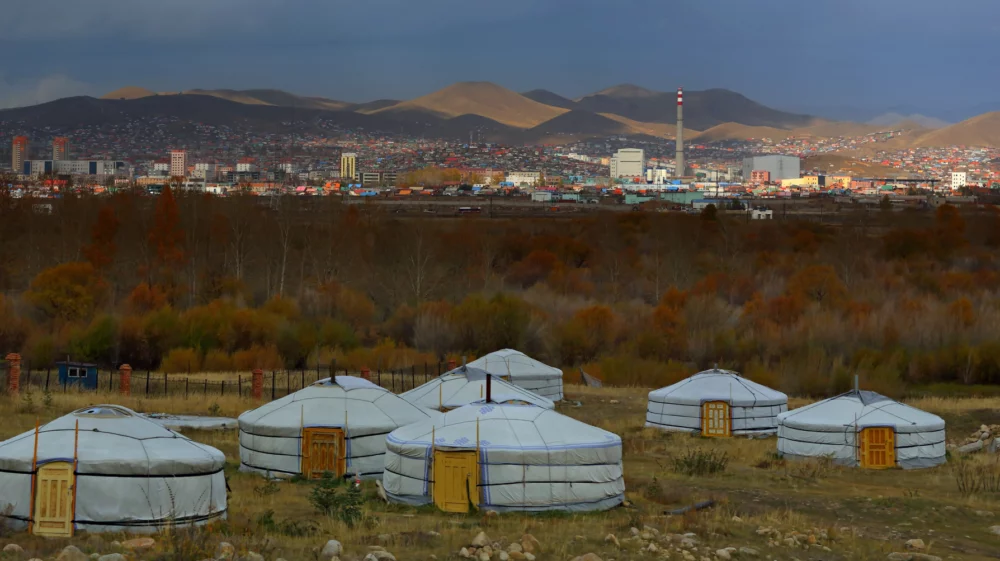 White yurts in the foreground, with a city and industrial chimney stacks behind, with a mountain range under a blue-gray cloudy sky.