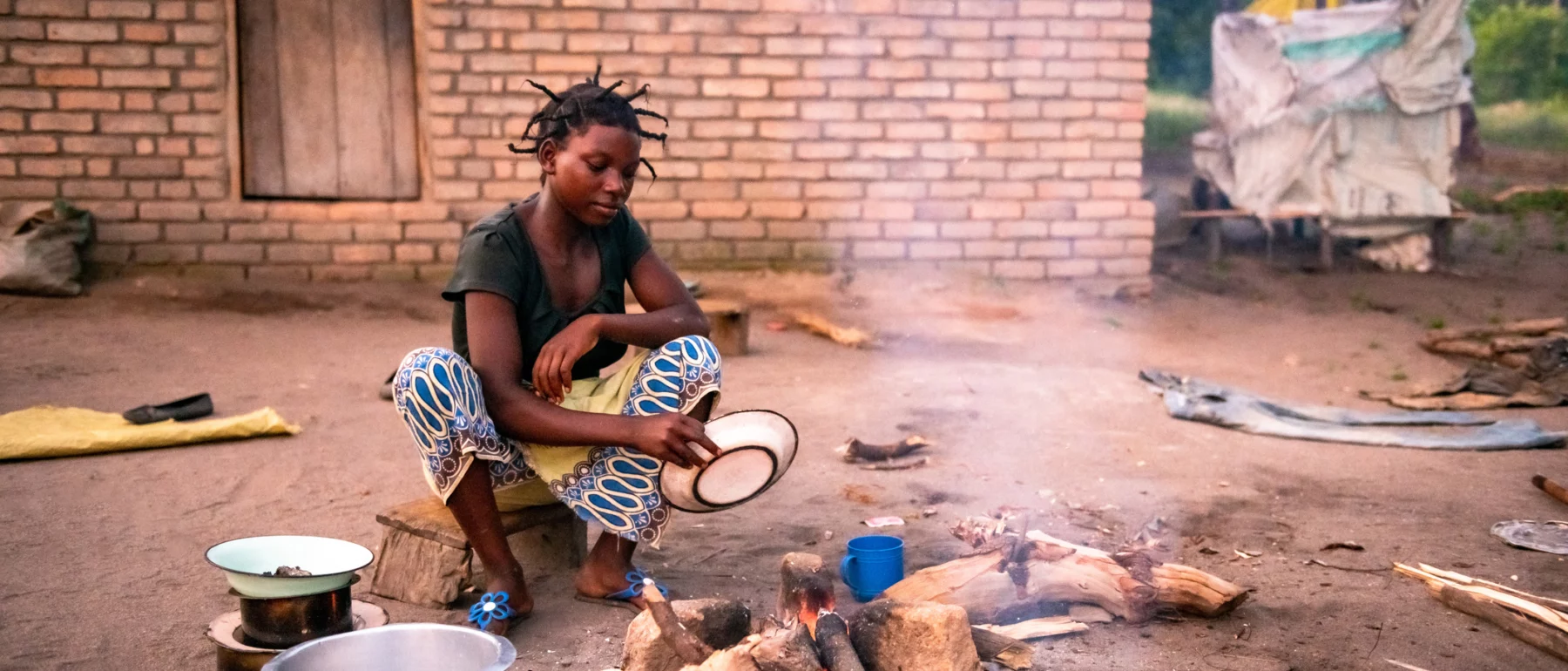 Young African woman cooks a meal over a wood-burning fire outside a brick house in Malawi