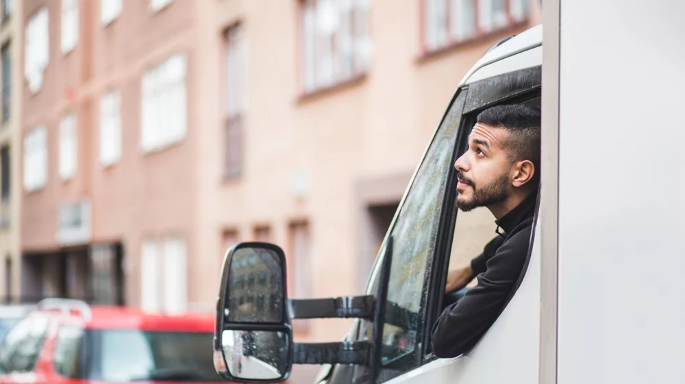 A transport worker leaning out of the window of a white van, looking to his side, with urban buildings in the background.