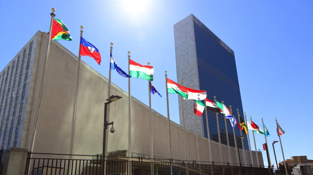 Flags of various countries flapping in front of the UN Headquarters in New York City with the sun shining in the background.
