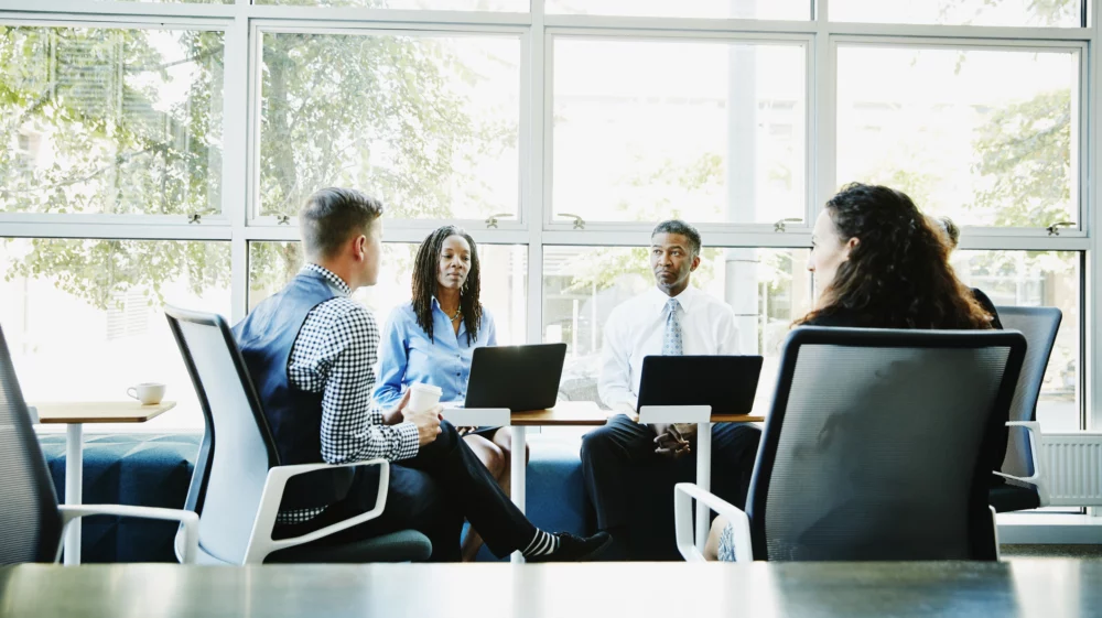 A group of diverse professionals engaged in a meeting around a table in a bright office setting with a large window and greenery outside.