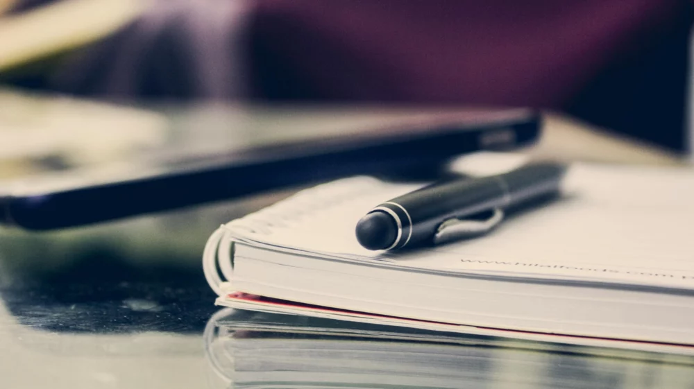 Open wire notebook with a black pen on top of it, lying on a glass table