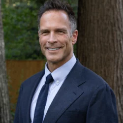 Ed Carr wears a navy suit and tie and smiles at the camera in an outdoor setting with a tree in the background