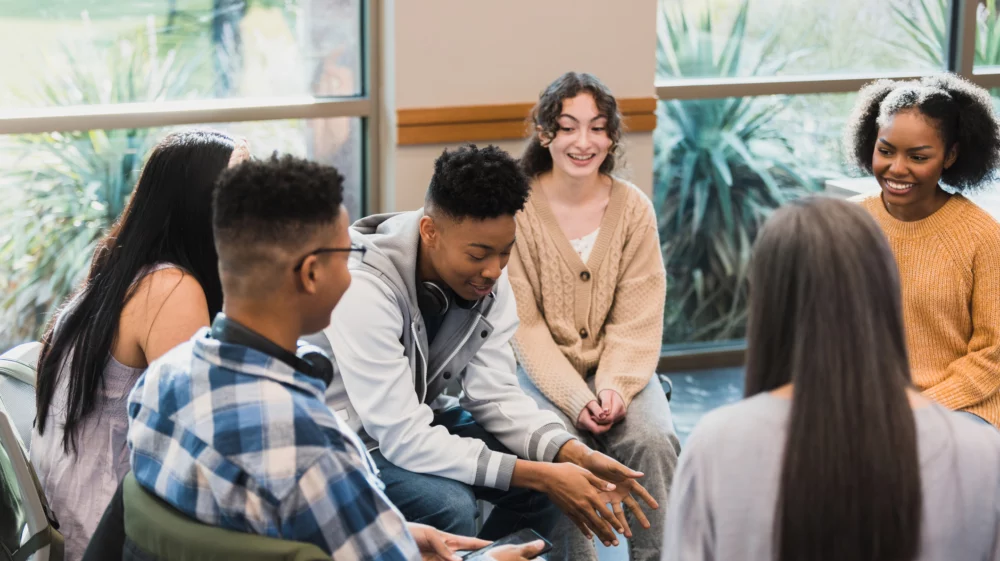 A seated group of six young people react to a young man in the centre of the group relaying a story.