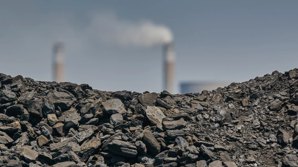 A close-up view of coal in the foreground with industrial smokestacks emitting white smoke in the blurry background, suggesting the theme of industrial pollution and fossil fuel emissions.