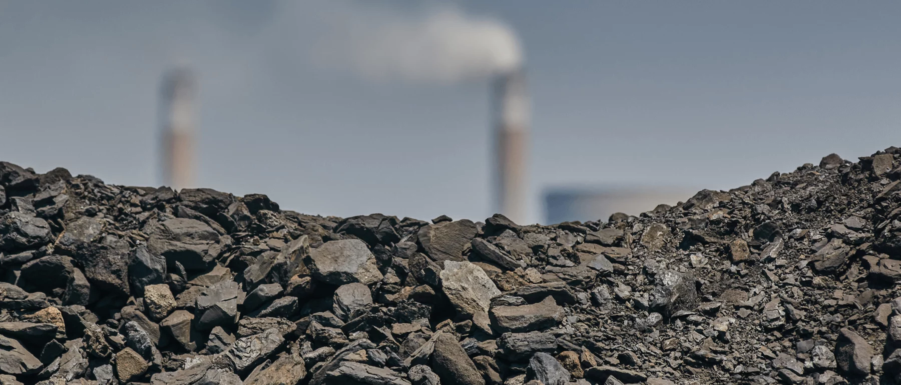 A close-up view of coal in the foreground with industrial smokestacks emitting white smoke in the blurry background, suggesting the theme of industrial pollution and fossil fuel emissions.