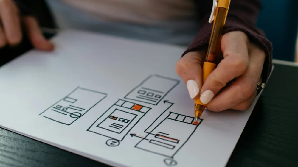 Close up of a woman's hand holding a pen, shading in a wireframe diagram. A wireframe is a 2D illustration of a web page's interface. Here, simple black-lined boxes are linked together with arrows.