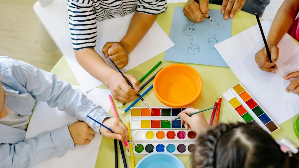 Aerial shot of four children and an adult sitting around a yellow table, painting.
