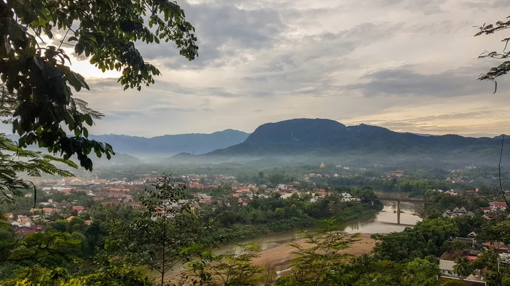 Aerial view of Mekong River winding through lush tropical trees and red-roofed buildings beneath gray dawn sky in Luang Prabang, Lao PDR.
