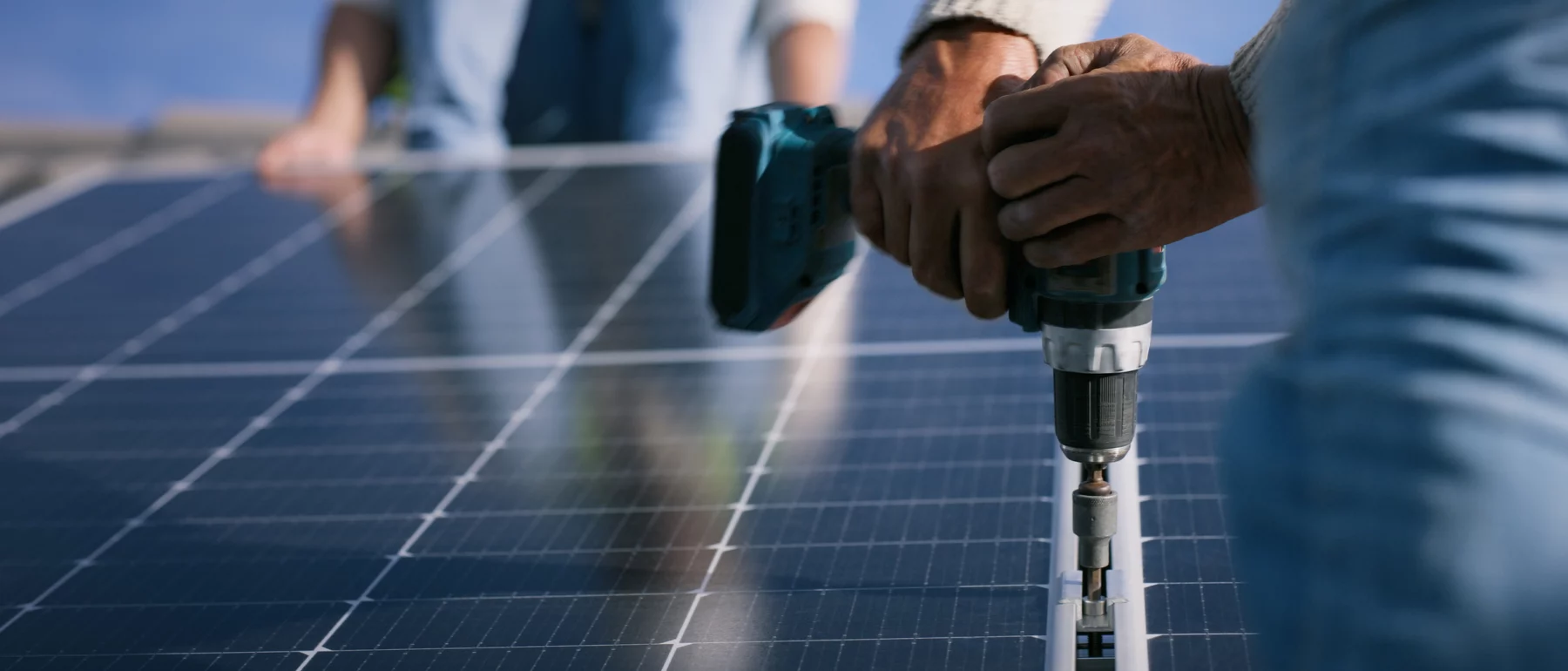 Technician hands using a cordless drill to secure a solar panel to a mounting structure on a clear day, reflecting the concept of renewable energy installations.
