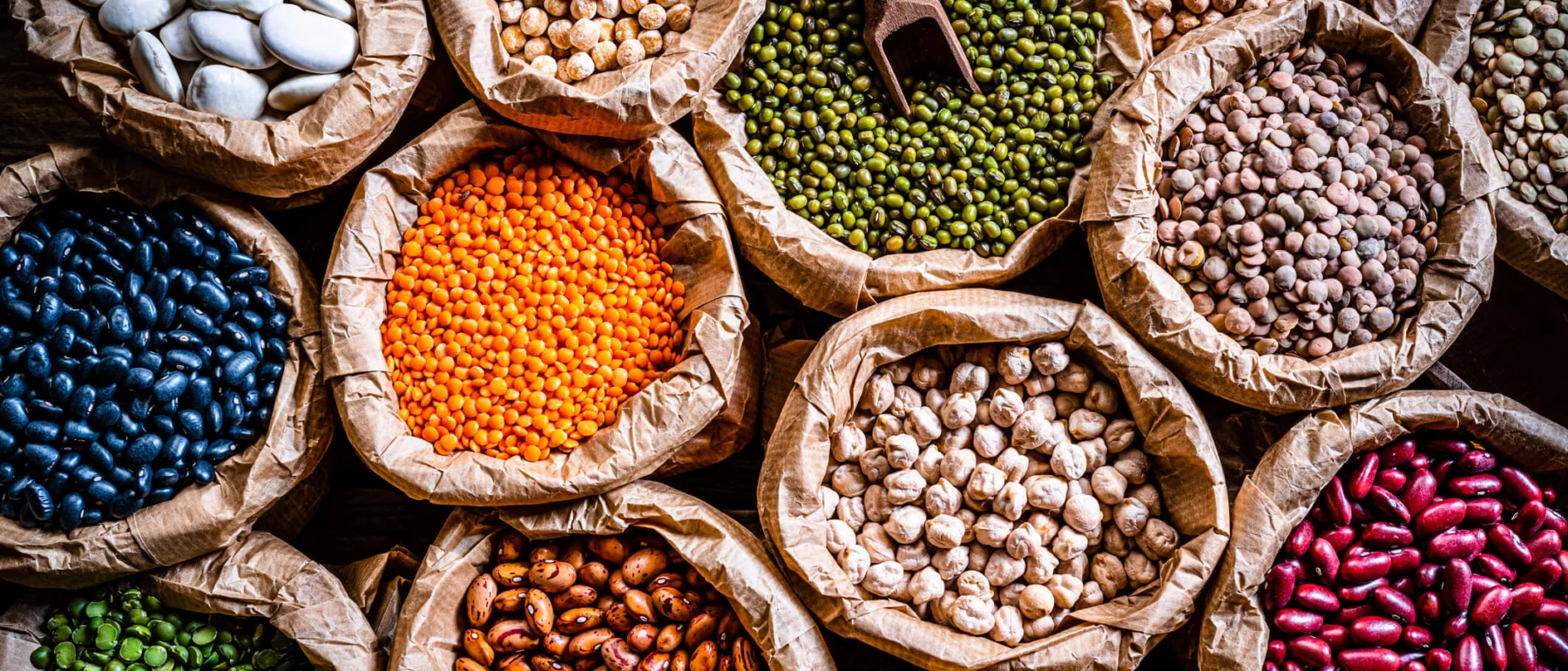 Dried beans and legumes of different colours in sacks, taken from above: black beans, green peas, red kidney beans, chickpeas, pinto beans, large white beans...