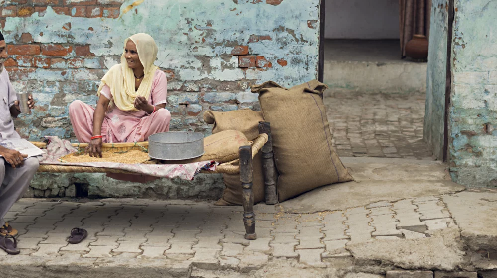 An older woman and an older man sitting on a bench in front of a blue painted wall smiling at each other.