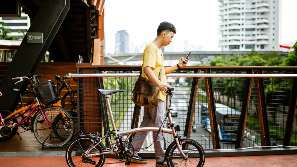 A cyclists walking with his bike on a pedestrian bridge while using his smartphone.