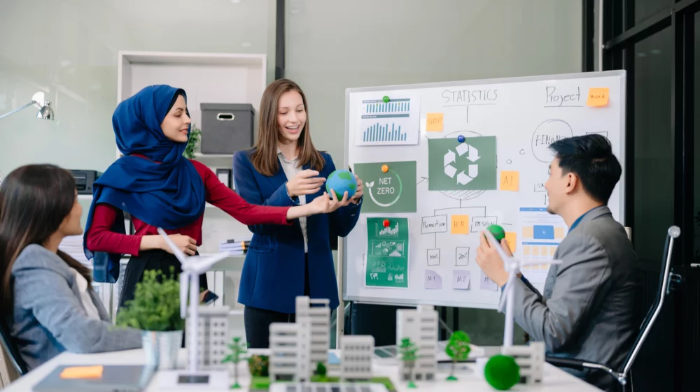 University students interacting with a model of part of a city, with a whiteboard in the background showing various images and text relating to sustainability and Net Zero.