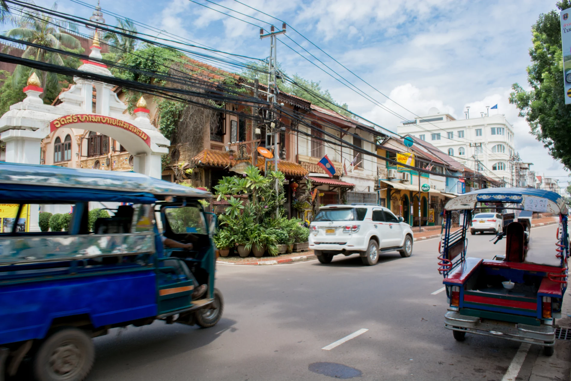 Two-storey buildings on one side of a two-lane road. Slightly blurred traffic rushes by away from the camera: a small white car and a blue tuk tuk.