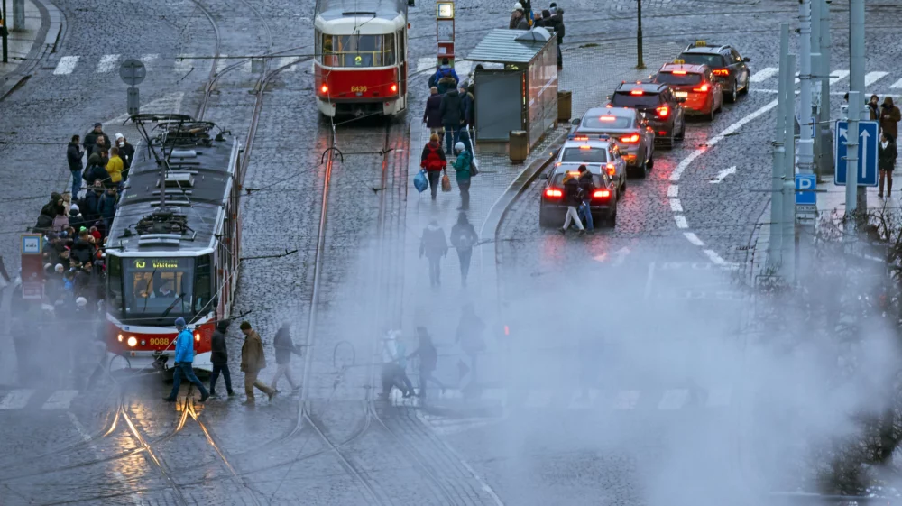 High angle shot of a street on a rainy day. Two red trams are on the left-hand side of the image, and a line of cars, brake lights visible, queues at a red traffic light on the right. Drifting smoke partially obscures the bottom half of the image. Pedestrians move around the space, crossing the road through the smoke and getting onto one of the trams.