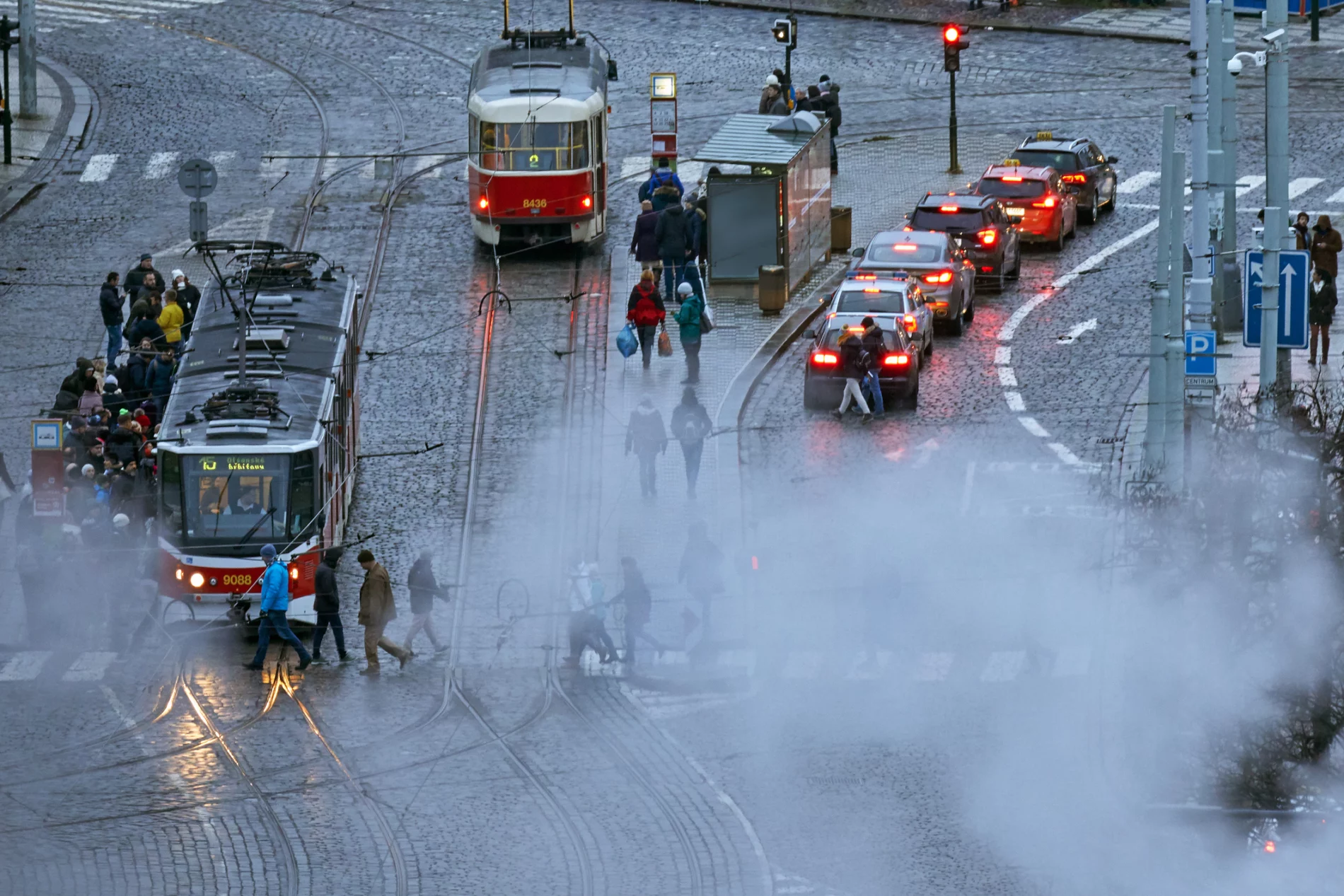 High angle shot of a street on a rainy day. Two red trams are on the left-hand side of the image, and a line of cars, brake lights visible, queues at a red traffic light on the right. Drifting smoke partially obscures the bottom half of the image. Pedestrians move around the space, crossing the road through the smoke and getting onto one of the trams.