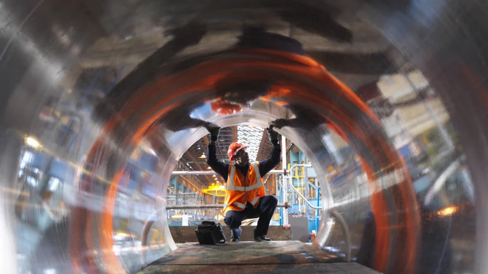 Worker inspecting the interior of a large metal pipe at an industrial site, with light reflecting off the curved surface creating a dynamic range of colors.