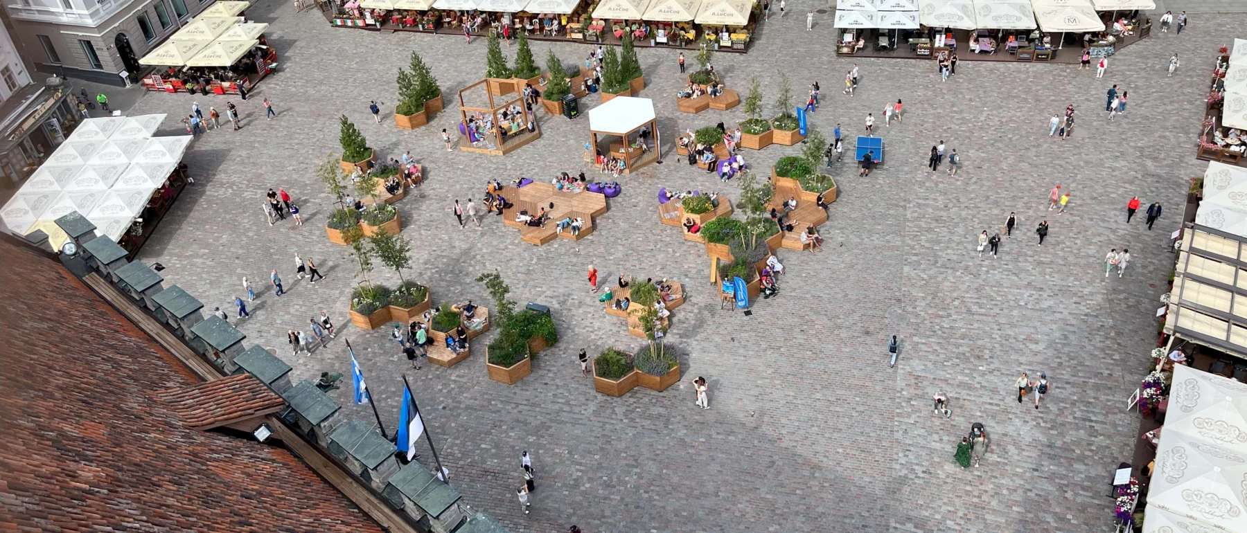 People gathered in a seating area in a city square edged by buildings in Tallinn, taken with a bird's eye view with an orange tile roof sloping away from the viewer's eye.