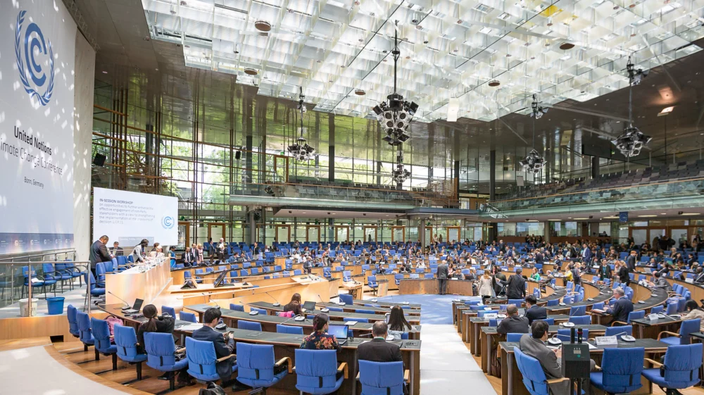 Panoramic view of a United Nations Climate Change Conference session held in a modern conference hall with floor-to-ceiling windows. Delegates from various countries are seated in a semi-circular arrangement, with a focus on a central presentation about enhancing stakeholder engagement for climate action. The large screen displays the session’s agenda, and the atmosphere is formal and attentive.