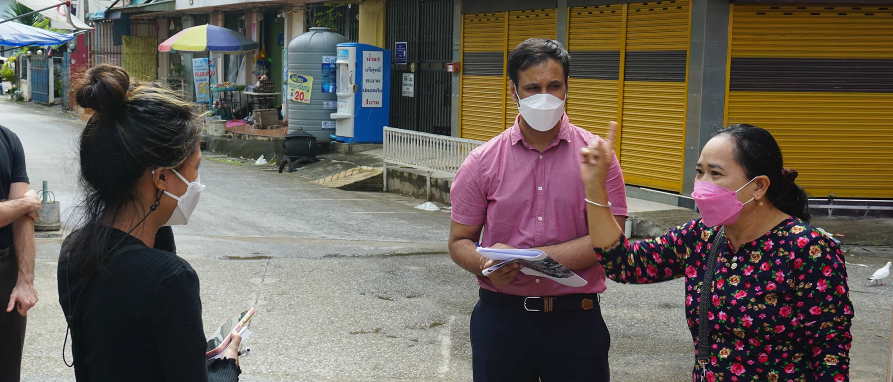 Three people standing on a street in Mae Sai, Thailand, wearing masks and having a conversation. One person, a woman in a floral shirt, is gesturing while speaking.