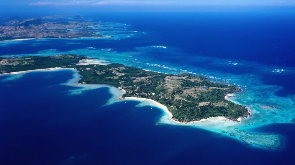 This image shows an aerial view of a tropical archipelago with numerous small islands and sandy beaches. The crystal-clear turquoise waters are vibrant and filled with coral reefs visible from above. The islands are interspersed with greenery and patches of forest, creating a striking contrast against the blue ocean. In the distance, the horizon meets a clear blue sky, enhancing the serene beauty of this coastal landscape.