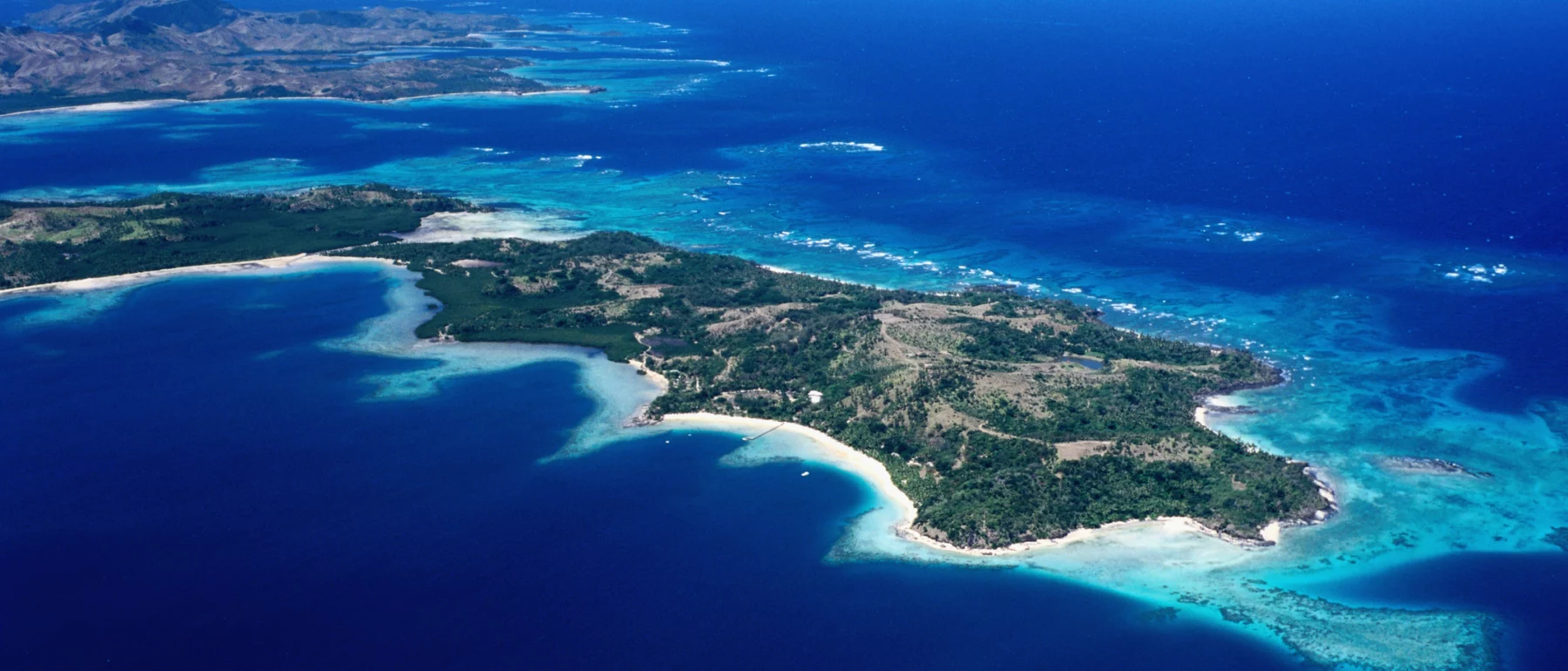 This image shows an aerial view of a tropical archipelago with numerous small islands and sandy beaches. The crystal-clear turquoise waters are vibrant and filled with coral reefs visible from above. The islands are interspersed with greenery and patches of forest, creating a striking contrast against the blue ocean. In the distance, the horizon meets a clear blue sky, enhancing the serene beauty of this coastal landscape.