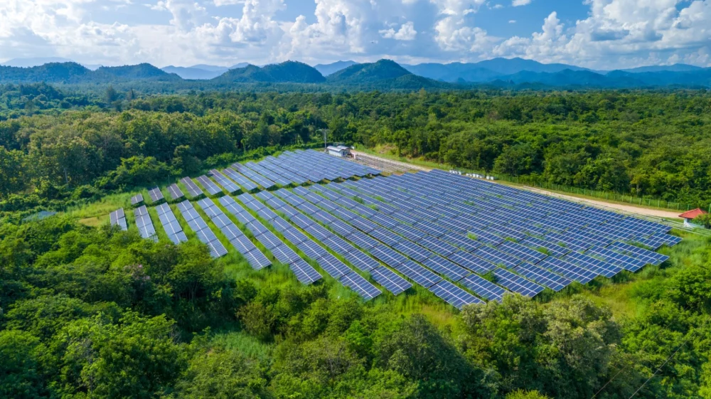 Aerial view of a large solar farm surrounded by lush green forests and mountains in the background under a partly cloudy sky.