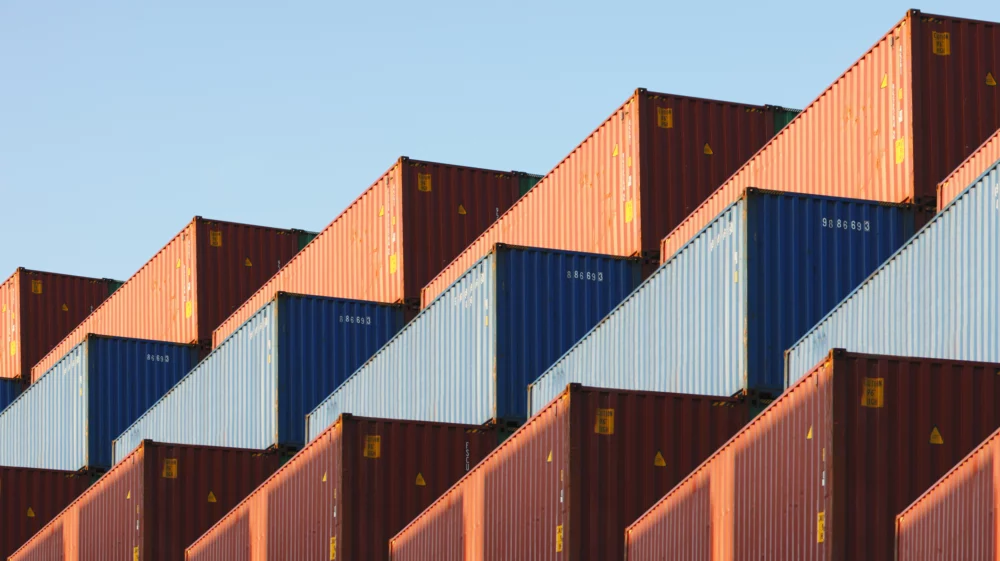 Stacked cargo containers at a shipping yard are arranged in a neat, diagonal pattern. The containers are primarily red and blue, set against a clear, pale blue sky. The arrangement creates a visually striking, geometric effect.