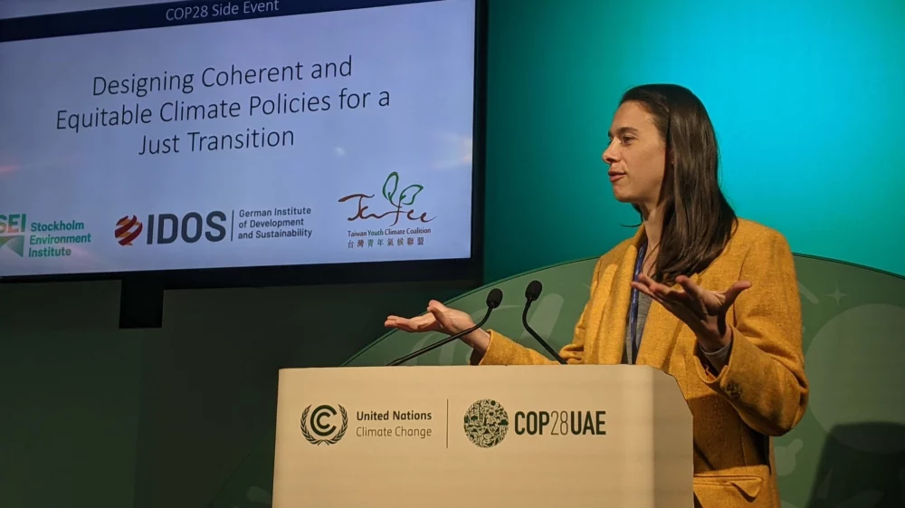 A woman stands at a lectern with a white screen and blue green background, hands raised at her shoulders, palms up.