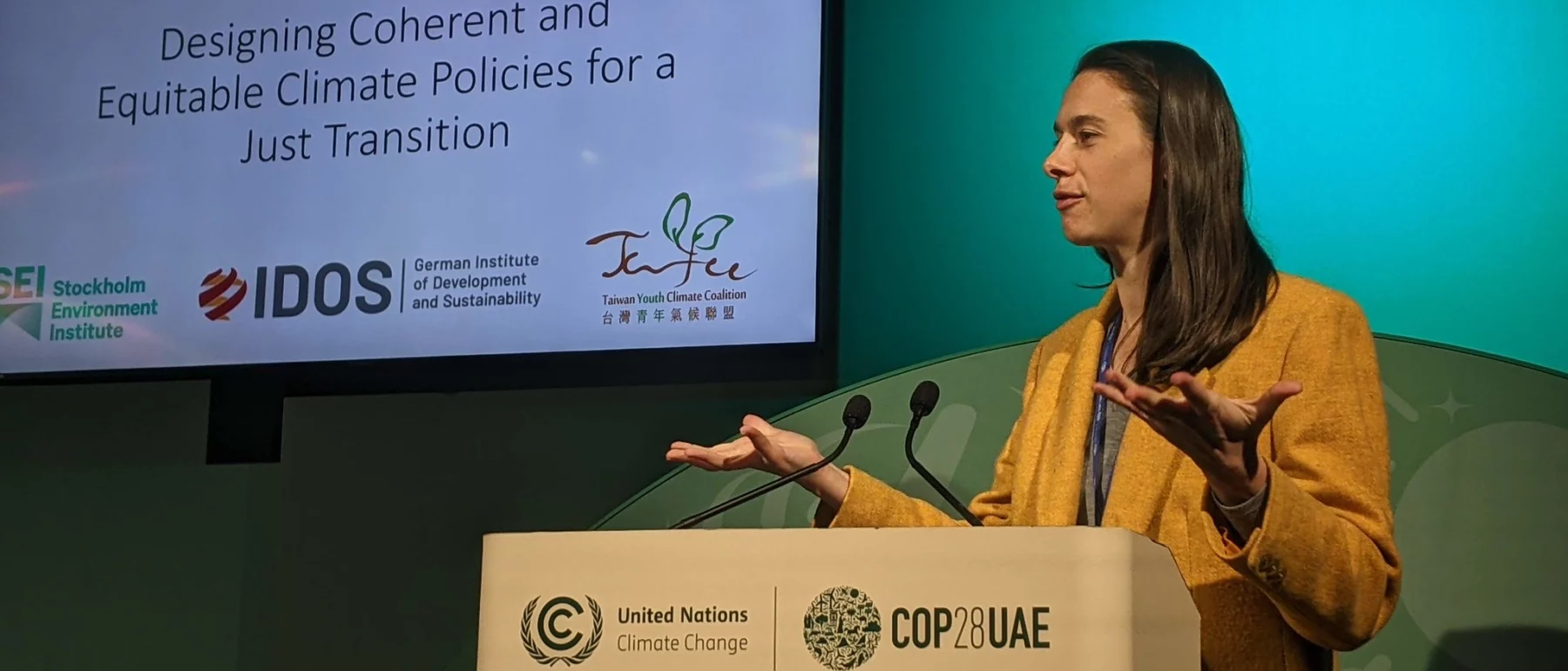 A woman stands at a lectern with a white screen and blue green background, hands raised at her shoulders, palms up.